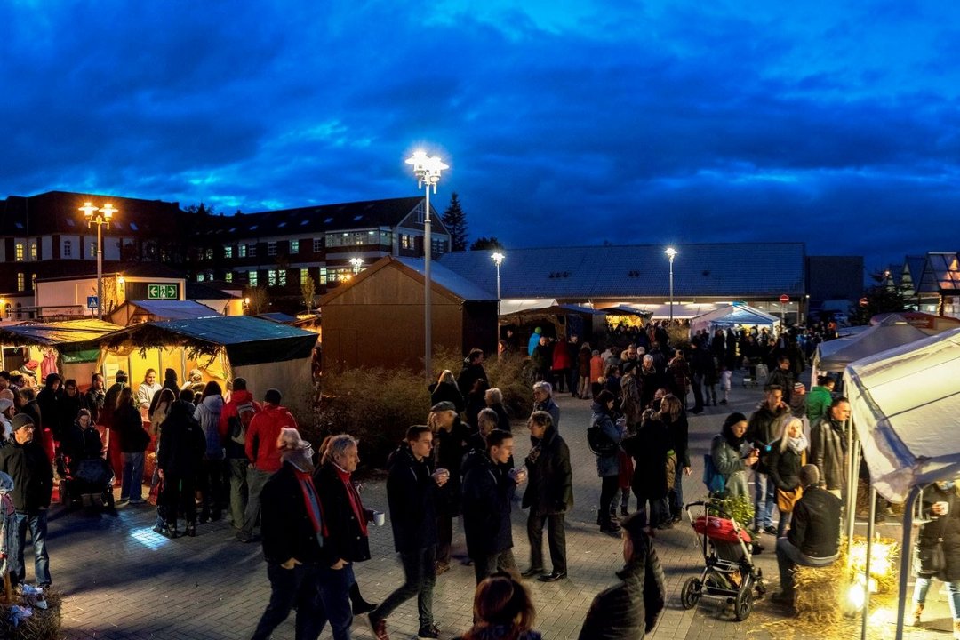 Unter nachtblauem Himmel ist ein Platz mit vielen erleuchteten Holzbuden und im Vordergrund ein erleuchtetes Zelt mit Stroh auf dem Boden zu sehen, an denen viele Besucher entlang gehen. 